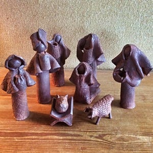 May include: A collection of handcrafted, dark brown ceramic figurines, including figures with draped cloaks, a seated figure, and a sheep. The figurines are arranged on a wooden surface, suggesting a nativity scene or similar theme.