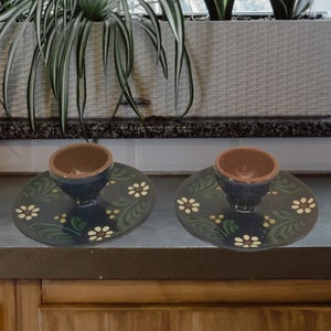 May include: Two blue ceramic egg cups with brown interiors, each sitting on a matching blue and white floral patterned saucer. The saucers have green leaves and white flowers.
