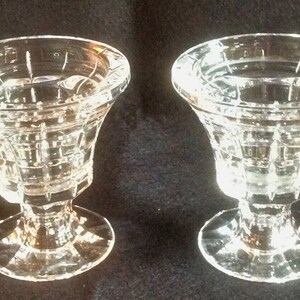 May include: Two clear glass candle holders featuring a flared rim and a geometric grid pattern on the exterior. Each piece stands on a circular base and has a thick stem connecting to the bowl, showcasing a faceted design that reflects light.