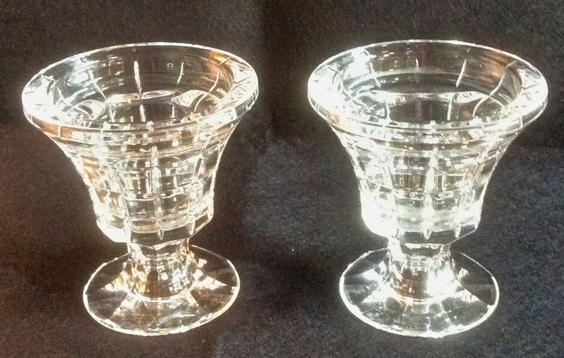 May include: Two clear glass candle holders featuring a flared rim and a geometric grid pattern on the exterior. Each piece stands on a circular base and has a thick stem connecting to the bowl, showcasing a faceted design that reflects light.