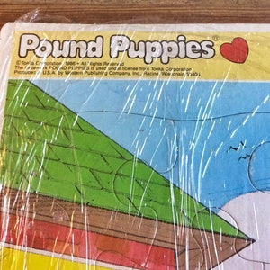 May include: A vintage Pound Puppies jigsaw puzzle box with a green roof, a red and yellow base, and a blue sky. The box is wrapped in plastic and has the text "Pound Puppies" and "Tonka Corporation 1986 All Rights Reserved" on it.