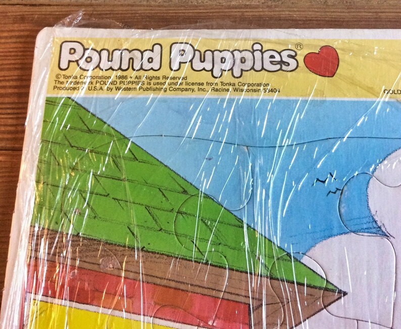 May include: A vintage Pound Puppies jigsaw puzzle box with a green roof, a red and yellow base, and a blue sky. The box is wrapped in plastic and has the text "Pound Puppies" and "Tonka Corporation 1986 All Rights Reserved" on it.