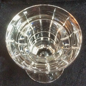 May include: A clear glass candle holder featuring a circular top opening with a stepped, tiered interior design. The exterior surface is adorned with a grid of vertical and horizontal cut lines, creating a series of raised rectangular panels.