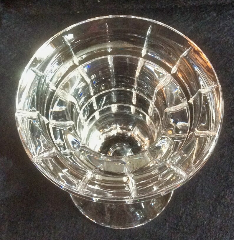 May include: A clear glass candle holder featuring a circular top opening with a stepped, tiered interior design. The exterior surface is adorned with a grid of vertical and horizontal cut lines, creating a series of raised rectangular panels.