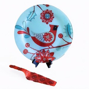 Può includere: Un piatto rotondo in vetro azzurro con un uccello cardinale rosso, motivi floreali rossi e foglie di agrifoglio color ottanio. Una spatola da portata in vetro rosso abbinata con un motivo floreale bianco è posta davanti su un supporto nero.