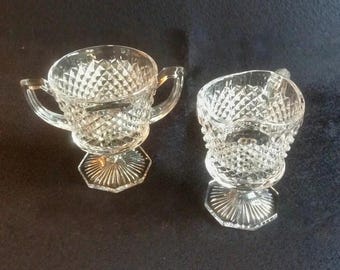 Op de afbeelding: Een suikerpot van helder glas met twee oren en een bijpassende melkkan, beide voorzien van een geperst ruitpatroon en een gefacetteerde zeshoekige voet. Het glaswerk heeft een getextureerd oppervlak en een transparante afwerking.