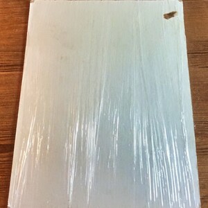 May include: A white, rectangular piece of cardboard wrapped in clear plastic wrap. The cardboard has a small, brown, rectangular piece of tape on the top right corner.
