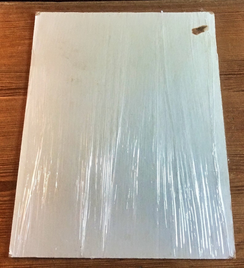 May include: A white, rectangular piece of cardboard wrapped in clear plastic wrap. The cardboard has a small, brown, rectangular piece of tape on the top right corner.