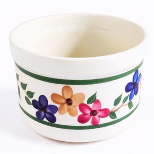May include: A round, off-white ceramic bowl featuring a horizontal green band near the top and bottom rim. The center section is decorated with a repeating pattern of five-petaled flowers in shades of blue, orange, and pink with green leaves.