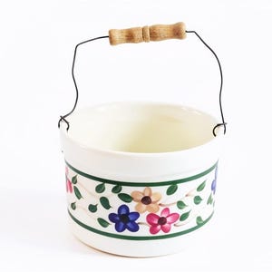 May include: A cream-colored ceramic bucket featuring a painted floral pattern with blue, pink, and tan flowers, green leaves, and dark green horizontal bands. The bucket includes a thin black wire handle with a central light brown wooden grip.