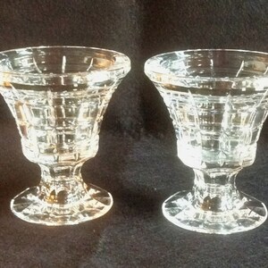 May include: Two clear glass pedestal cups featuring a geometric grid pattern on the exterior. Each cup has a wide, flared rim, a short stem, and a circular base with a faceted edge, reflecting light through the transparent glass construction.