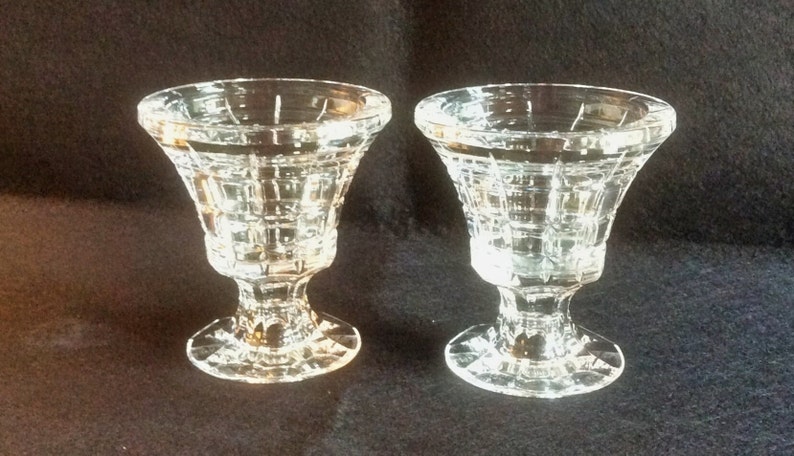 May include: Two clear glass pedestal cups featuring a geometric grid pattern on the exterior. Each cup has a wide, flared rim, a short stem, and a circular base with a faceted edge, reflecting light through the transparent glass construction.