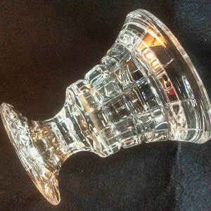 May include: A clear glass pedestal bowl featuring a geometric grid pattern of raised squares across its exterior surface. The vessel has a wide, flared rim and a sturdy, thick base, with a transparent finish that reflects light through its faceted design.