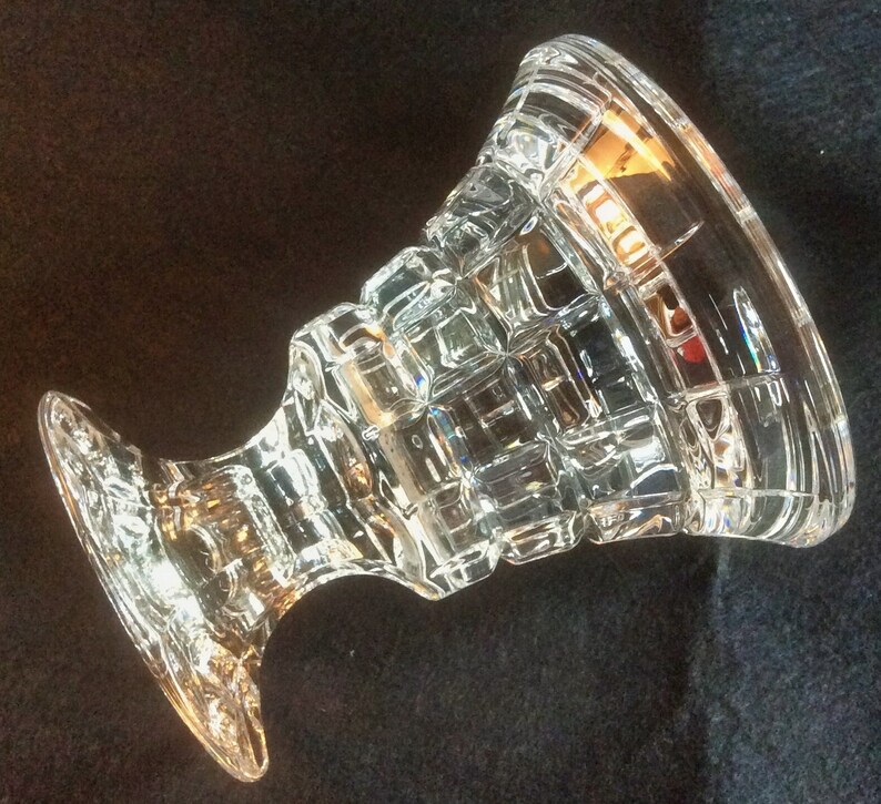 May include: A clear glass pedestal bowl featuring a geometric grid pattern of raised squares across its exterior surface. The vessel has a wide, flared rim and a sturdy, thick base, with a transparent finish that reflects light through its faceted design.