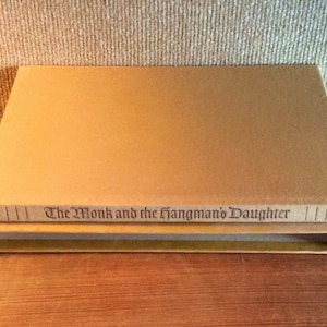 May include: A brown hardcover book with the title "The Monk and the Hangman's Daughter" printed on the spine.