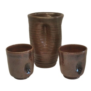 May include: A set of three brown ceramic cups, including a larger jug and two smaller cups. The cups have a textured, speckled finish and a unique, slightly asymmetrical shape.