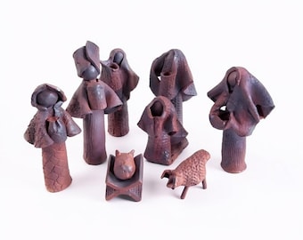 Puede incluir: Un conjunto de ocho figuras de arcilla de color marrón rojizo oscuro que representan un belén. La colección incluye varias figuras de pie con prendas drapeadas, una figura pequeña en una cuna rectangular y una figura de animal.