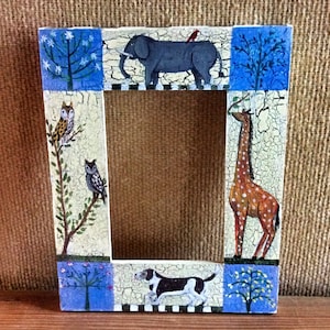 May include: A white wooden picture frame with a distressed finish. The frame is decorated with painted images of animals, including an elephant, a giraffe, a dog, and owls. The frame has a blue, white, and green colour scheme.