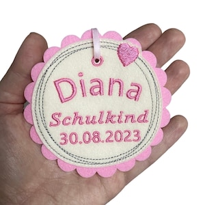 Könnte beinhalten: Rosa Filz-Kreis mit gewellten Rändern und einem weißen Zentrum. Das Zentrum hat einen grauen Kreis mit dem Text "Diana Schulkind 30.08.2023" in rosa Stickgarn gestickt. Ein kleines rosa Herz ist in der oberen rechten Ecke gestickt.