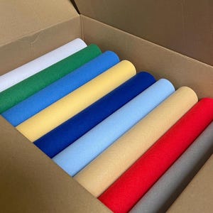 Op de afbeelding: Een doos gevuld met rollen viltstof in verschillende kleuren, waaronder wit, groen, blauw, geel, lichtblauw, beige, rood en grijs.