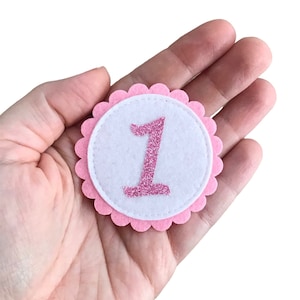 Könnte beinhalten: Rosa Filz Kreis mit einem weißen Zentrum und einer pinkfarbenen Glitzerzahl eins.