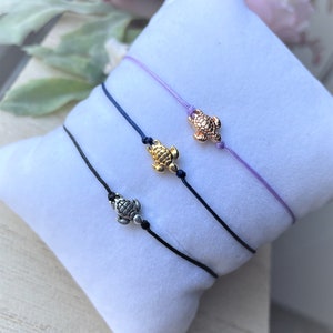 Könnte beinhalten: Drei verstellbare Armbänder aus Schnur mit einem kleinen Schildkrötenanhänger. Die Armbänder sind in Schwarz, Blau und Lila.