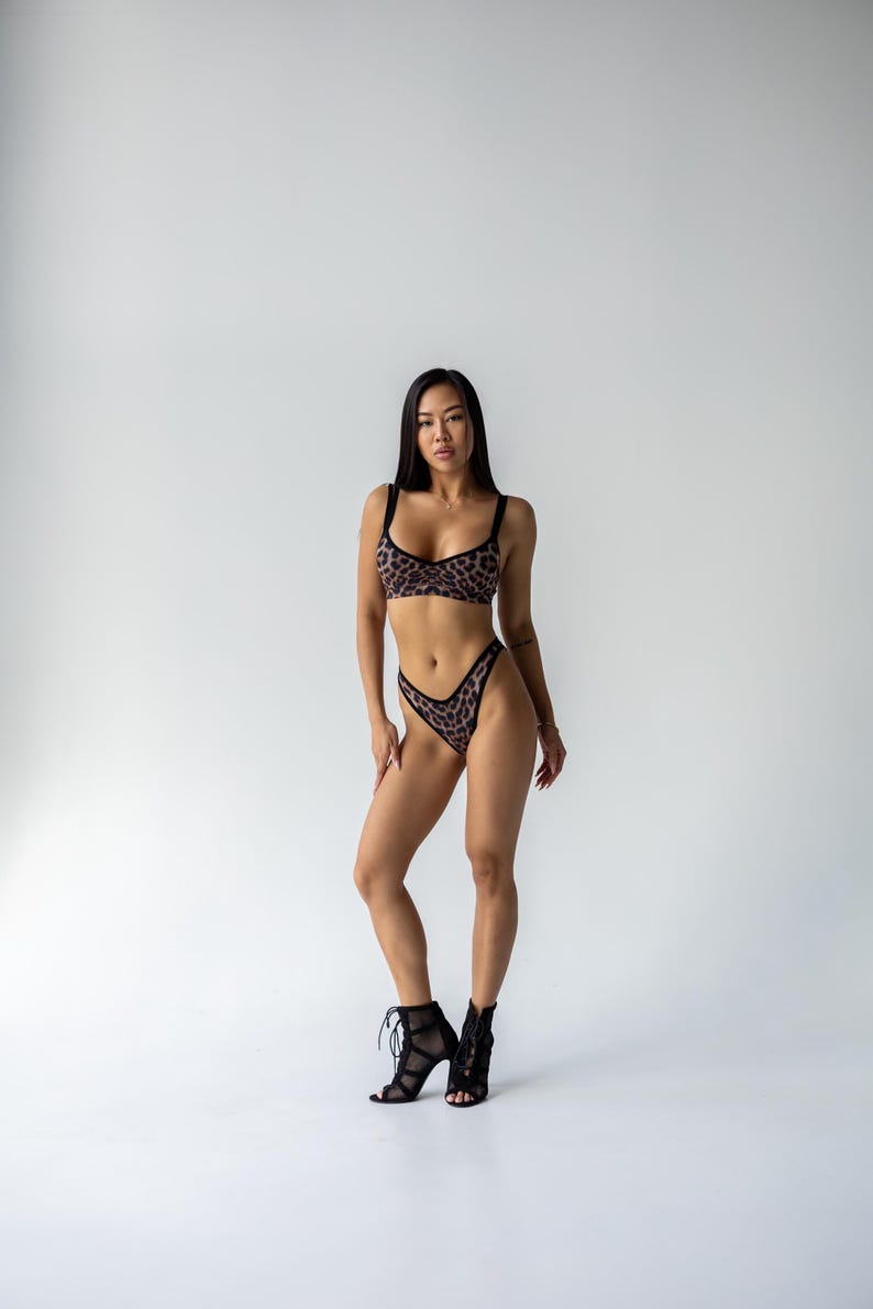 May include: A two-piece leopard print lingerie set with a matching bra and high-cut bottoms. The bra has black straps and trim. The model is wearing black lace-up heels. The background is plain white.
