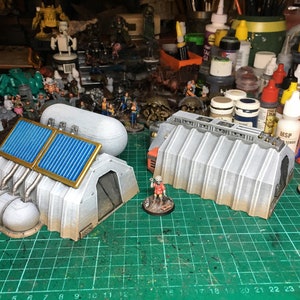May include: Two grey, corrugated metal buildings with a tunnel entrance. One building has two blue and gold solar panels on top. The buildings are on a green cutting mat.