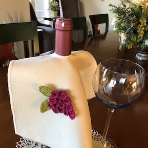 Puede incluir: Una bolsa de vino de lino blanco con un diseño de uva de ganchillo. La bolsa está drapeada sobre una botella de vino y una copa de vino está sentada sobre un encaje de ganchillo.