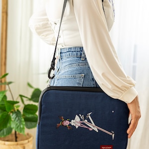May include: A navy blue laptop sleeve featuring a cartoon illustration of a girl with red hair and a white dress. The sleeve has a black strap and a small white label with the text "Oliver & Tanzen Design" on it.