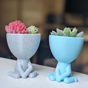 May include: Two 3D printed succulent planters, one gray and one light blue, shaped like a person sitting cross-legged.