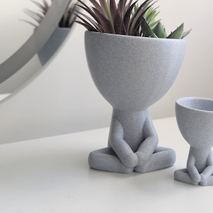 Puede incluir: Dos macetas de cerámica gris con forma de personas sentadas en posición de meditación. La maceta más grande tiene una planta verde con hojas en su interior.