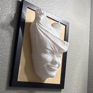 May include: A white plaster sculpture of a face with fabric draped over the head, mounted in a black frame on a light gray wall.