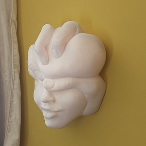 May include: A white wall sculpture depicting a human face with a hand covering the eyes. The sculpture is mounted on a yellow wall, with a white curtain visible on the left side of the image. The sculpture has a smooth, matte finish.
