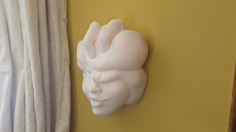 May include: A white wall sculpture depicting a human face with a hand covering the eyes. The sculpture is mounted on a yellow wall, with a white curtain visible on the left side of the image. The sculpture has a smooth, matte finish.