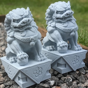 以下が含まれることがあります： 複雑なディテールが施された灰色の石製のフー・ドッグ像2体。花柄の正方形の台座の上に座っています。像は正面を向いており、口を開けて舌を出しています。