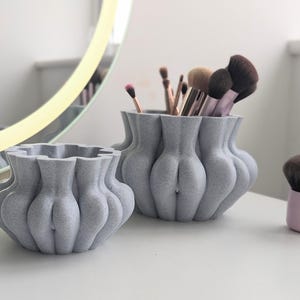 Könnte beinhalten: Zwei graue Keramikvasen mit einer strukturierten, gewellten Oberfläche. Die größere Vase ist mit Make-up-Pinseln gefüllt. Die kleinere Vase ist leer.