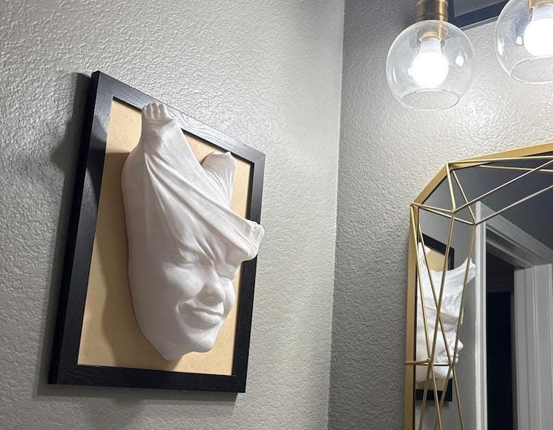 May include: A white plaster sculpture of a face with fabric draped over the head, mounted in a black frame on a light gray wall.