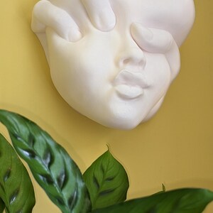 May include: A white plaster wall sculpture of a face with a hand covering the eyes. The sculpture is mounted on a yellow wall, with green plant leaves in the foreground.