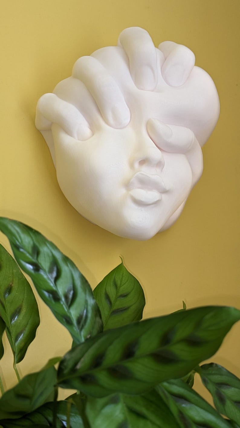 May include: A white plaster wall sculpture of a face with a hand covering the eyes. The sculpture is mounted on a yellow wall, with green plant leaves in the foreground.