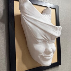 May include: A white plaster sculpture of a face with a cloth draped over the top of the head. The face is smiling and the cloth is draped over the head in a way that makes it look like the person is trying to hide their face.