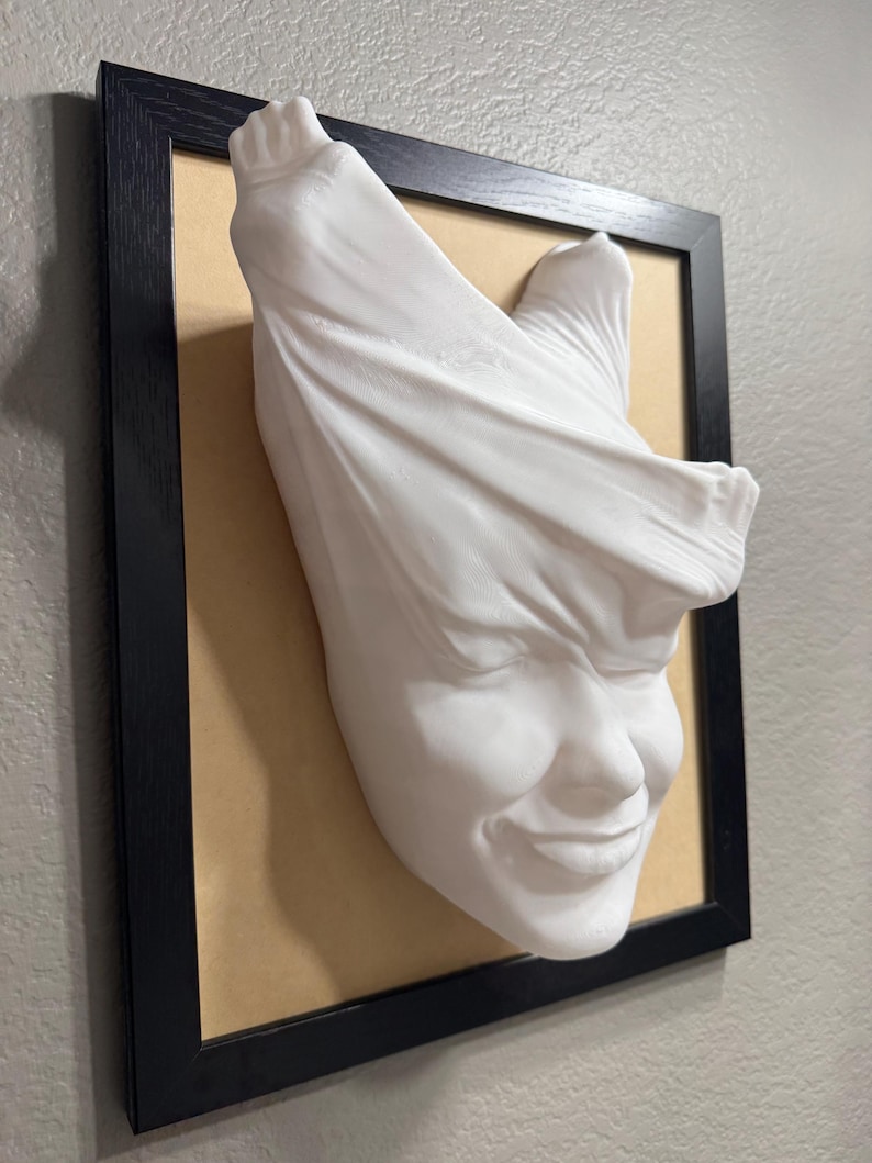 May include: A white plaster sculpture of a face with a cloth draped over the top of the head. The face is smiling and the cloth is draped over the head in a way that makes it look like the person is trying to hide their face.