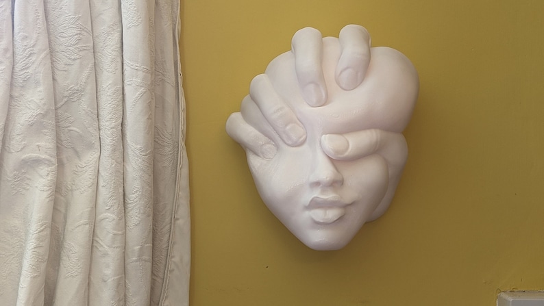 May include: A white plaster wall sculpture of a face with hands covering the eyes. The sculpture is mounted on a yellow wall, with a white curtain visible on the left side.
