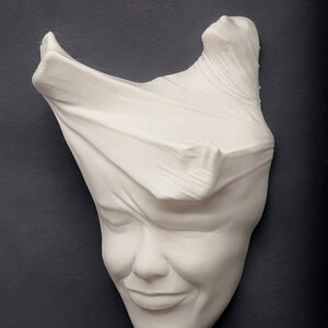 May include: White plaster sculpture of a face with a cloth draped over the top of the head. The face is smiling.