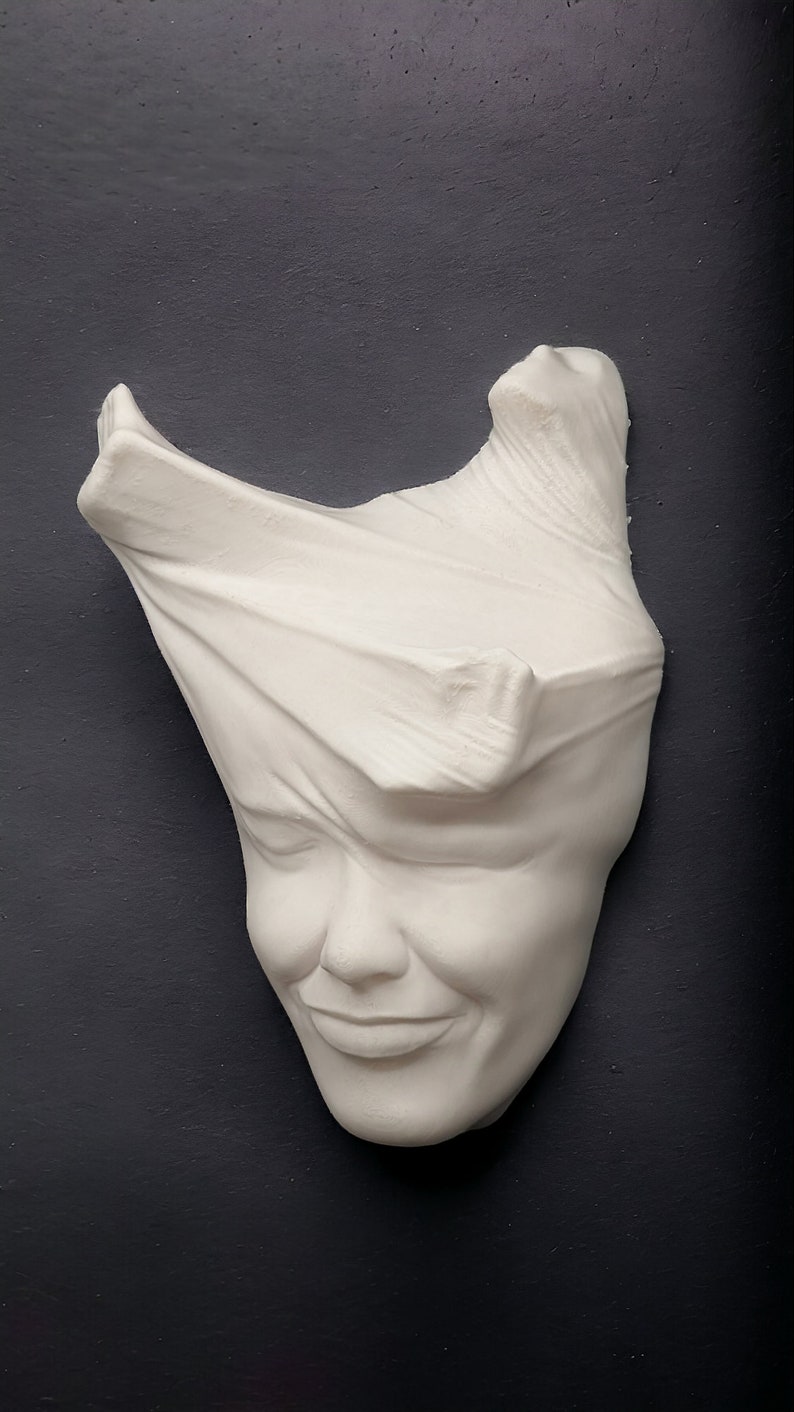 May include: White plaster sculpture of a face with a cloth draped over the top of the head. The face is smiling.