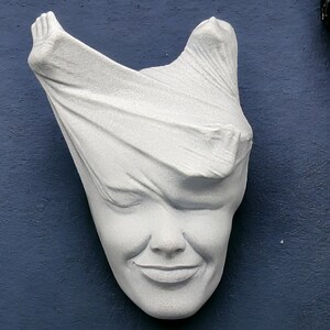 May include: A white plaster sculpture of a face partially covered by a cloth. The face is smiling.