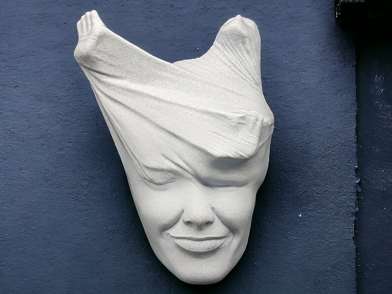 May include: A white plaster sculpture of a face partially covered by a cloth. The face is smiling.