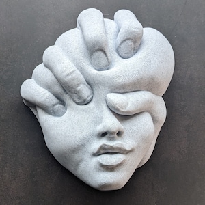 May include: A gray sculpture of a human face with hands covering the eyes and part of the face. The sculpture has a textured surface and is set against a dark background. The artwork is a unique piece of home decor.