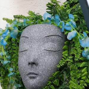 Può includere: Un vaso di pietra grigia a forma di viso con gli occhi chiusi e un'espressione neutra. Il vaso è circondato da piante verdi frondose e fiori blu.