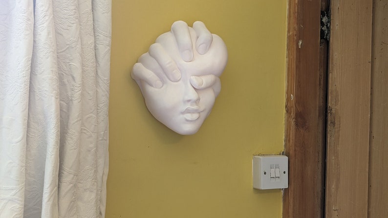 May include: A white plaster wall sculpture of a face with hands covering the eyes. The sculpture is mounted on a yellow wall. A white curtain is to the left of the sculpture.